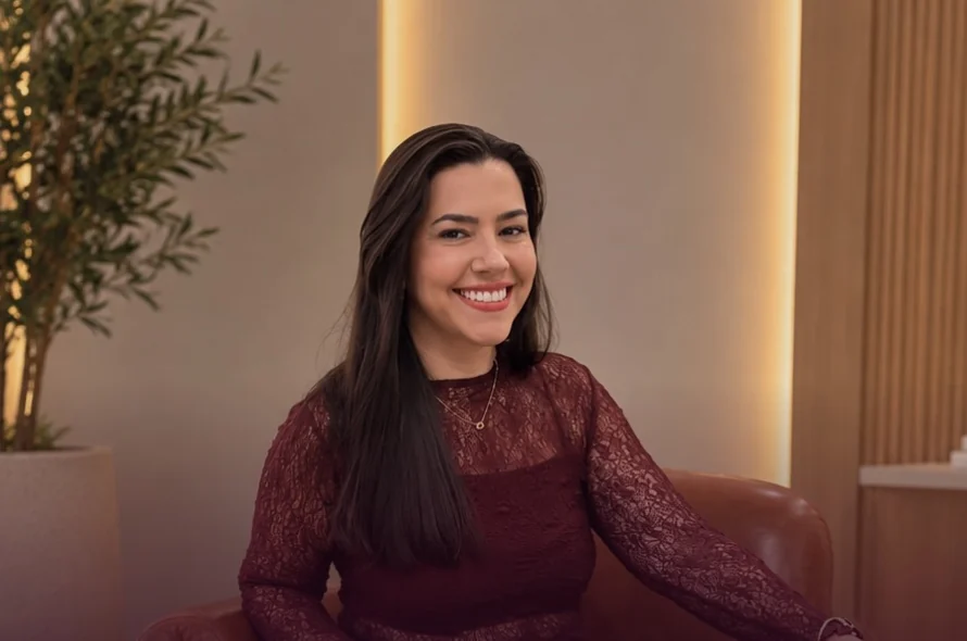 Jaque sorrindo numa poltrona de couro, numa sala com plantas, parede iluminada e painel de madeira